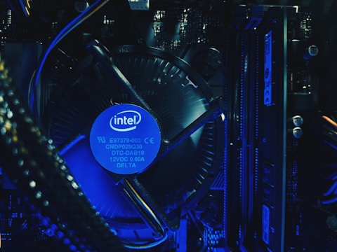 A close-up view of a computer's internal hardware components, including a central processing unit (CPU) cooler with a prominently displayed Intel logo. The lighting has a blue tone, highlighting the intricacy of the various cables, circuit boards, and heat sinks in the assembly.