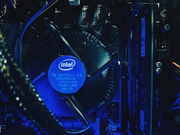 A close-up view of a computer's internal hardware components, including a central processing unit (CPU) cooler with a prominently displayed Intel logo. The lighting has a blue tone, highlighting the intricacy of the various cables, circuit boards, and heat sinks in the assembly.