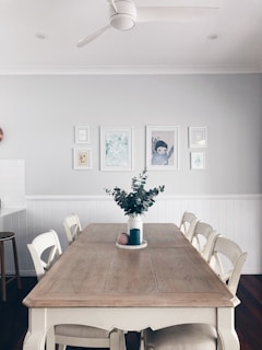 A dining room with a large wooden table surrounded by six white chairs. The table features a blue vase with green foliage and a round pink object. The walls are painted light grey and decorated with a series of framed artworks. A ceiling fan is mounted on the white ceiling, which has recessed lighting.