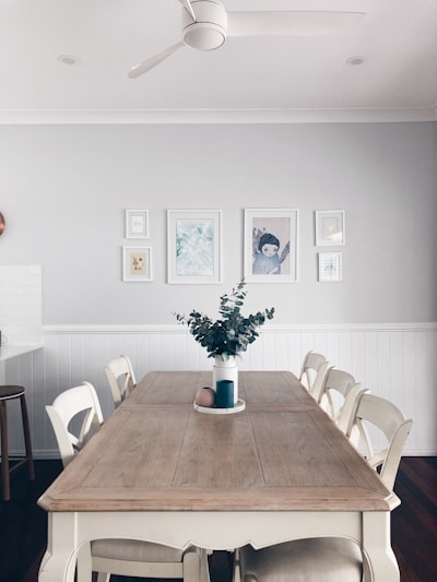 A dining room with a large wooden table surrounded by six white chairs. The table features a blue vase with green foliage and a round pink object. The walls are painted light grey and decorated with a series of framed artworks. A ceiling fan is mounted on the white ceiling, which has recessed lighting.