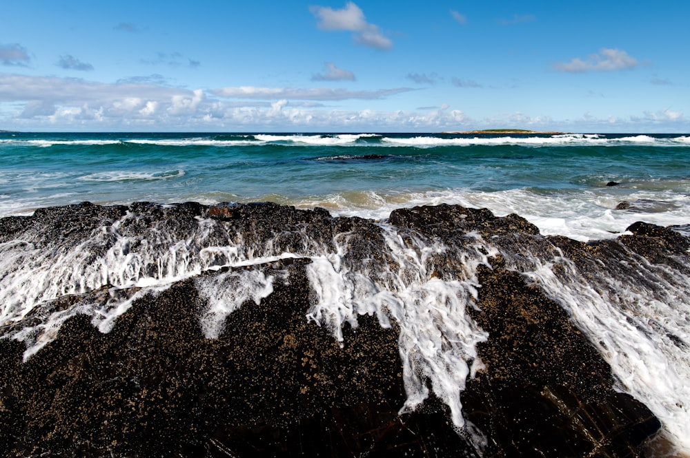 ocean waves crashing on black rock formation under blue sky during daytime