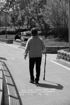 A person walks down a paved path using a walking stick. Trees and bushes line the path, creating a natural setting. In the background, there are two people sitting on a low wall, appearing engaged in conversation. The image is in black and white, enhancing the contrast and shadows.