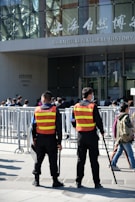Two security personnel wearing reflective vests stand in front of a crowd, near barricades, outside the Shanghai Natural History Museum. The scene is busy with many people around, and the building's glass facade and signage are visible.