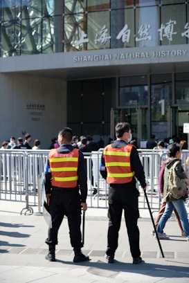 Two security personnel wearing reflective vests stand in front of a crowd, near barricades, outside the Shanghai Natural History Museum. The scene is busy with many people around, and the building's glass facade and signage are visible.
