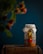 A rustic jar filled with a colorful mix of ingredients sits atop a wooden surface. The jar is sealed with a fabric cover tied with twine. In the foreground, there's a blurred hint of foliage with red and green colors. The background is a deep blue, adding contrast to the vibrant contents of the jar.