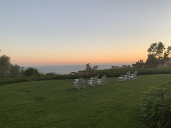 A peaceful grassy landscape with several white lounge chairs facing an ocean view. The horizon is softly illuminated by a sunset, presenting a gradual transition from orange to blue. Trees and bushes surround the area, adding to the serene atmosphere.