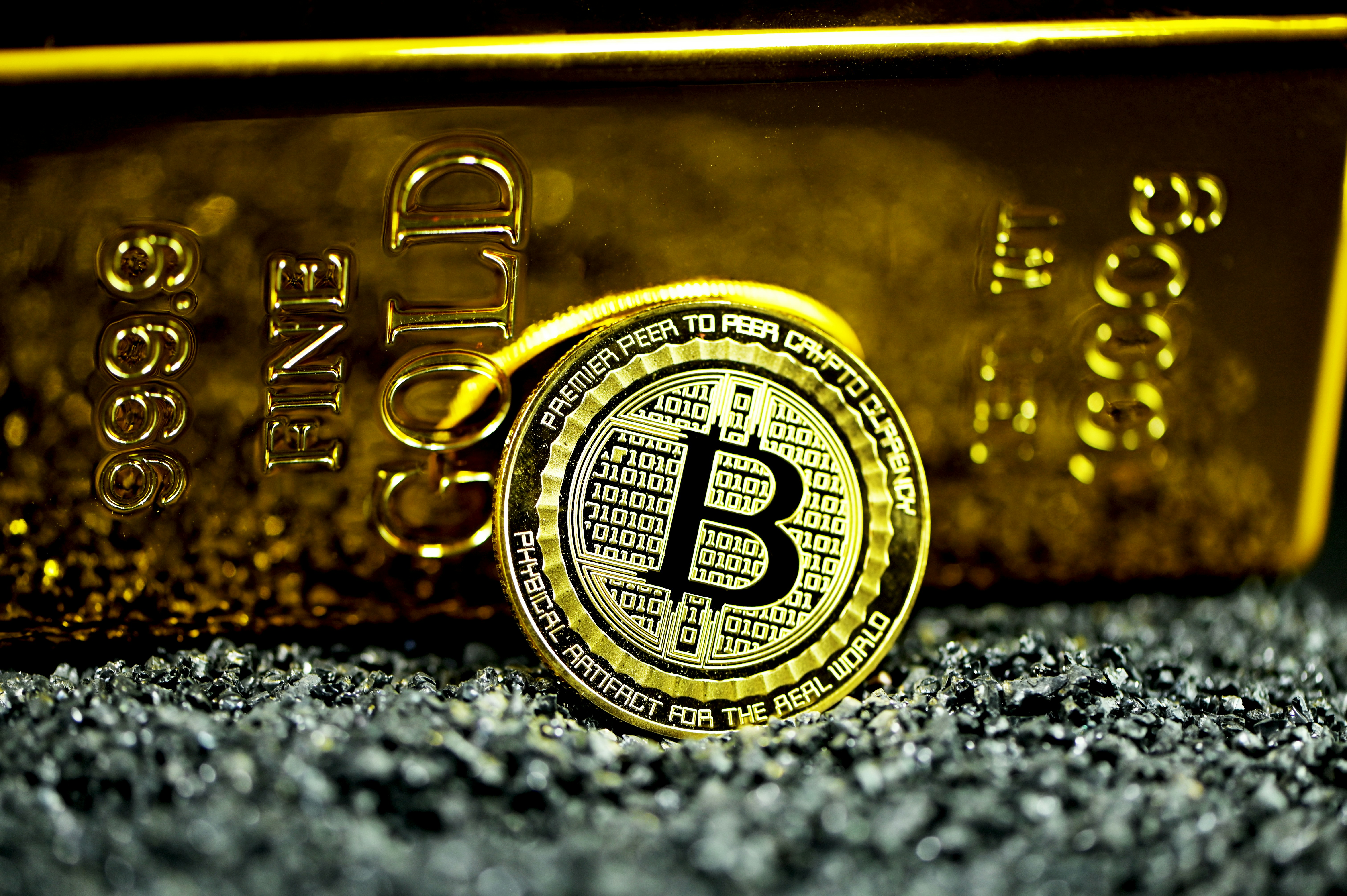 Bitcoin propped up against gold bullion on black stones.