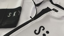 A close-up of a white and black sports jacket featuring a visible zipper. The fabric has a textured, mesh-like pattern. The words 'SKY' and 'SKYFLY' are printed on the jacket in black letters.