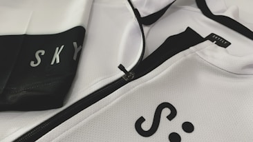 A close-up of a white and black sports jacket featuring a visible zipper. The fabric has a textured, mesh-like pattern. The words 'SKY' and 'SKYFLY' are printed on the jacket in black letters.
