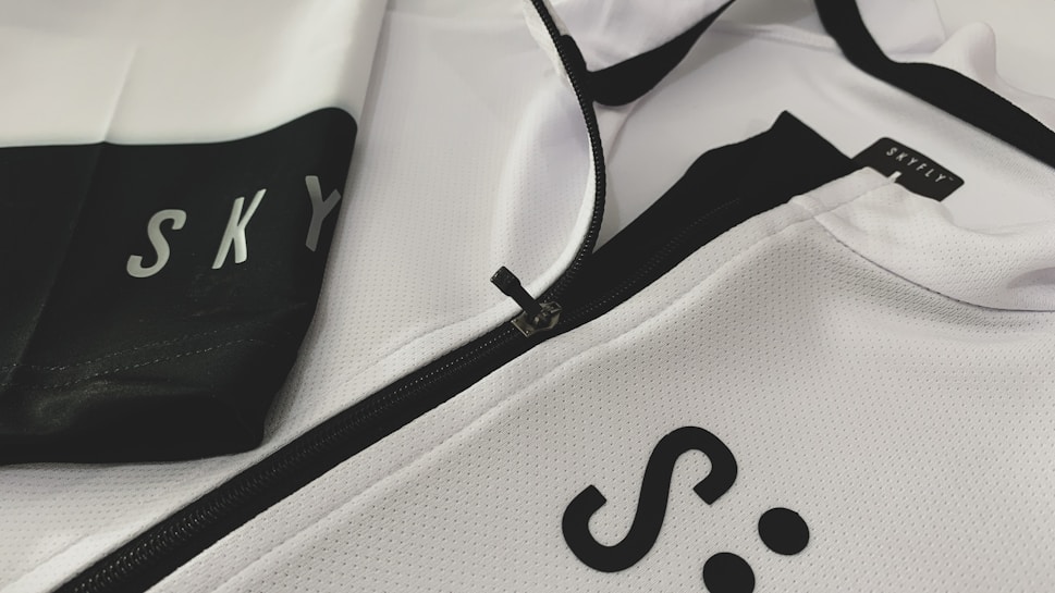A close-up of a white and black sports jacket featuring a visible zipper. The fabric has a textured, mesh-like pattern. The words 'SKY' and 'SKYFLY' are printed on the jacket in black letters.