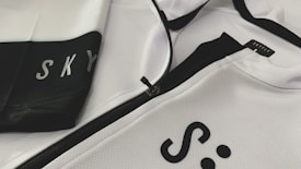 A close-up of a white and black sports jacket featuring a visible zipper. The fabric has a textured, mesh-like pattern. The words 'SKY' and 'SKYFLY' are printed on the jacket in black letters.