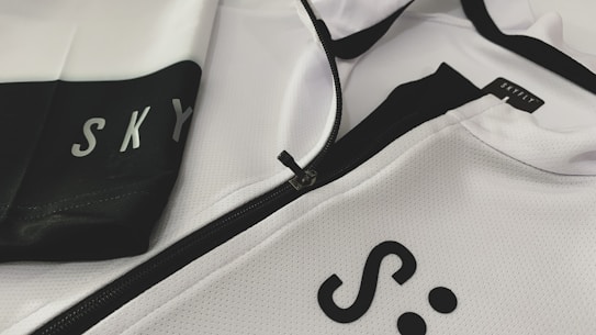 A close-up of a white and black sports jacket featuring a visible zipper. The fabric has a textured, mesh-like pattern. The words 'SKY' and 'SKYFLY' are printed on the jacket in black letters.