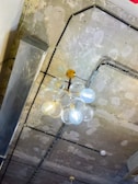 A modern light fixture is hanging from an industrial-style ceiling. The fixture consists of clear glass globes clustered together around a golden central rod. The ceiling has an unfinished, rough texture with visible patches and exposed wiring.