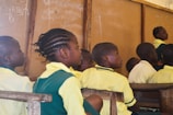 A group of children in school uniforms sit attentively in a classroom setting. The wooden desks and a chalkboard with writing in the background suggest a typical classroom environment. The children appear focused on someone or something outside the frame.