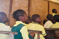 A group of children in school uniforms sit attentively in a classroom setting. The wooden desks and a chalkboard with writing in the background suggest a typical classroom environment. The children appear focused on someone or something outside the frame.
