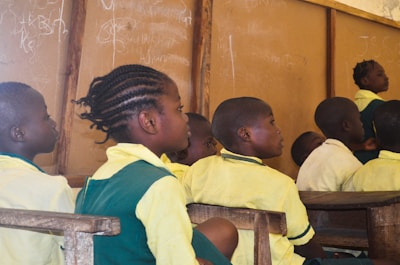 A group of children in school uniforms sit attentively in a classroom setting. The wooden desks and a chalkboard with writing in the background suggest a typical classroom environment. The children appear focused on someone or something outside the frame.