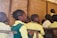 A group of children in school uniforms sit attentively in a classroom setting. The wooden desks and a chalkboard with writing in the background suggest a typical classroom environment. The children appear focused on someone or something outside the frame.
