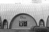 A modern architectural building with a textured, arched design features a large, prominent window display of the Dior brand. The exterior is characterized by a repetitive pattern on the facade, and the landscape includes neatly trimmed bushes in the foreground.
