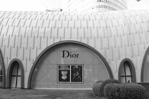 A modern architectural building with a textured, arched design features a large, prominent window display of the Dior brand. The exterior is characterized by a repetitive pattern on the facade, and the landscape includes neatly trimmed bushes in the foreground.