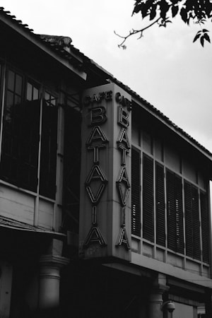 A black and white image featuring a tall, vertical sign attached to the corner of a building. The sign reads 'Cafe Batavia' in bold, illuminated letters. The building has large windows with shutters and is topped with a tile roof. The scene captures a sense of architectural character.