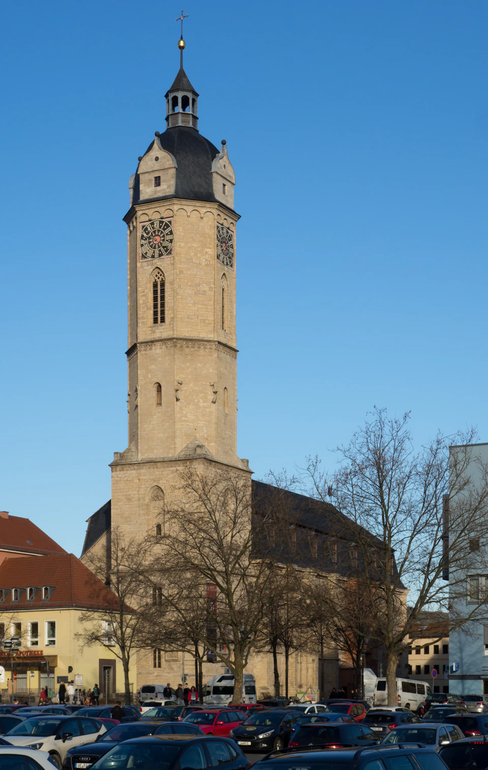 The image depicts a tall, light-colored stone church with a clock tower reaching towards the clear blue sky. The church is situated on a street corner and features two clocks - one on each side of the tower. In front of the church, there are several cars parked along the street, including a few red cars that stand out against the otherwise monochrome scene.

In addition to the church and cars, there are also people present in the image. Some individuals can be seen walking around or standing near the church, while others are scattered throughout the scene. The overall atmosphere of the image conveys a sense of tranquility and calmness, with the church as the focal point against the backdrop of the blue sky.