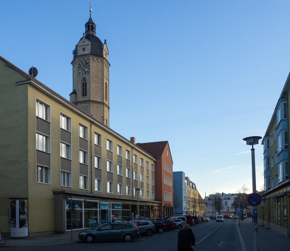 The image depicts a street scene with a tall, ornate clock tower on the right side and a row of buildings lining both sides of the street. The sky above is clear blue, suggesting a bright and sunny day. On the left side of the street, there are several cars parked along the curb, including one car that appears to be in motion as it passes by.

A person can be seen walking on the sidewalk near the center of the image, adding life to the scene. The overall atmosphere is calm and peaceful, with a sense of order and organization evident in the well-maintained street and buildings lining both sides of the road.