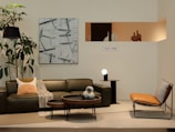 A modern living room setup featuring a dark green leather sofa with decorative pillows and a blanket, a small side table with a unique white and black striped vase, and two round coffee tables. A contemporary artwork hangs on the wall behind the sofa, and a large plant with broad leaves stands to the left. A leather lounge chair with a checkered cushion is positioned to the right. A minimalist niche displaying abstract sculptures is visible on the wall.