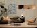 A modern living room setup featuring a dark green leather sofa with decorative pillows and a blanket, a small side table with a unique white and black striped vase, and two round coffee tables. A contemporary artwork hangs on the wall behind the sofa, and a large plant with broad leaves stands to the left. A leather lounge chair with a checkered cushion is positioned to the right. A minimalist niche displaying abstract sculptures is visible on the wall.