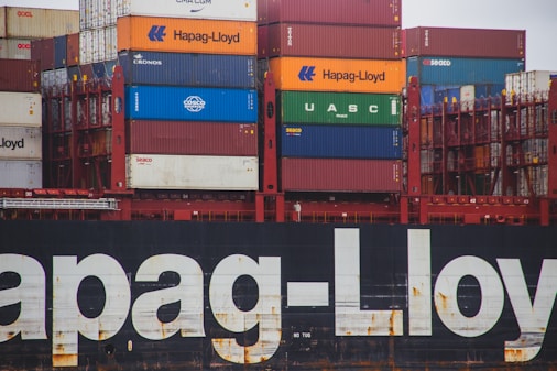 A stack of colorful shipping containers on a cargo ship, featuring prominent brands like Hapag-Lloyd, UASC, and COSCO. The containers are organized in neat rows and the hull of the ship is visible with large branding painted on it.