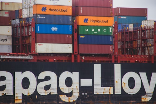 A stack of colorful shipping containers on a cargo ship, featuring prominent brands like Hapag-Lloyd, UASC, and COSCO. The containers are organized in neat rows and the hull of the ship is visible with large branding painted on it.