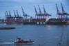 A large cargo ship marked with 'MSC' is docked at a port, with several towering cranes lined up along the dock. In the foreground, a smaller red passenger boat is moving across the water. The sky is overcast, and there are large wind turbines in the background.