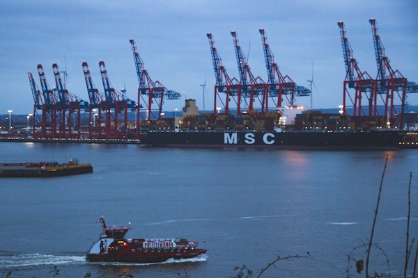 A large cargo ship marked with 'MSC' is docked at a port, with several towering cranes lined up along the dock. In the foreground, a smaller red passenger boat is moving across the water. The sky is overcast, and there are large wind turbines in the background.