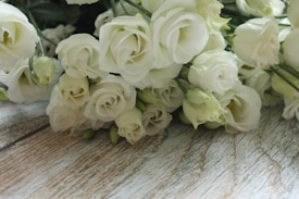 A bouquet of white roses arranged on a wooden surface. The flowers appear fresh and delicate, with soft petals and slight hints of green in the stems and buds. The wooden surface has a rustic texture, adding a natural backdrop to the elegant flowers.