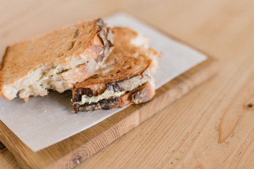 A grilled cheese sandwich placed on a piece of parchment paper atop a wooden cutting board. The sandwich appears to have been partially eaten, revealing the melted cheese inside. The surface is a light wooden table.
