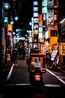 A vibrant street scene at night with a delivery scooter in the foreground. The vehicle is branded with KFC delivery logos and is traveling down a narrow alley lined with numerous illuminated signs in various colors. The ambiance is bustling and lively, with people walking along the sidewalk.
