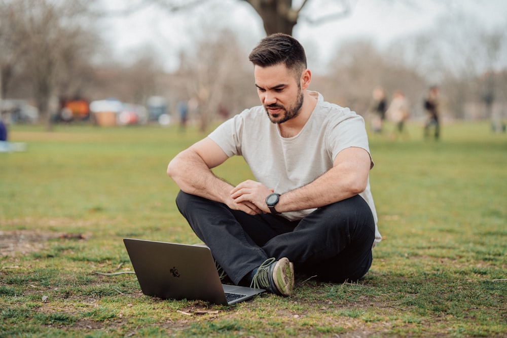 man in white crew neck t-shirt and black pants sitting on green grass field using