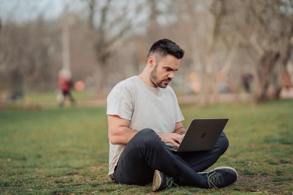 man in white crew neck t-shirt and black pants sitting on green grass field using