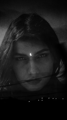 A monochrome image featuring a woman's face with long hair, looking directly at the viewer. A small crescent moon symbol is positioned between her eyebrows, and the background is dark, adding a mysterious or ethereal quality.