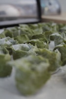 Green, uncooked dumplings arranged on a flat surface, likely in a kitchen setting. The perspective focuses on the texture and shape of the dumplings, which are sprinkled with flour, giving them a slightly frosted appearance.