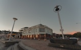 A large, multi-story building with the word 'HYDRO' written on it, surrounded by street lamps and situated in a paved urban area. The architecture features large windows and a flat roof. The scene includes a few cars parked on the side, and the sky is clear with a hint of sunset colors in the background.
