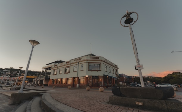 A large, multi-story building with the word 'HYDRO' written on it, surrounded by street lamps and situated in a paved urban area. The architecture features large windows and a flat roof. The scene includes a few cars parked on the side, and the sky is clear with a hint of sunset colors in the background.