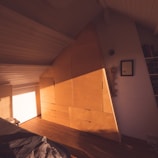 The image depicts an attic room with sloped ceiling, featuring a large, triangular wooden wardrobe built into the wall. The room is bathed in warm, natural light, creating a cozy atmosphere. On the right side, there is a small framed picture and a shelf filled with books.