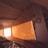 The image depicts an attic room with sloped ceiling, featuring a large, triangular wooden wardrobe built into the wall. The room is bathed in warm, natural light, creating a cozy atmosphere. On the right side, there is a small framed picture and a shelf filled with books.