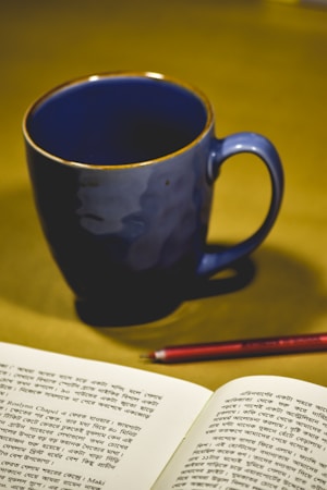 A blue ceramic mug sits on a surface next to an open book with printed text in Bengali script. A red pencil is placed between the mug and the book, suggesting a setting of study or contemplation.