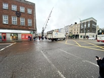 A city street intersection with several buildings, including a store with a large sale sign advertising a 60% discount. A truck is taking a turn in the middle of the intersection, and there are pedestrians on the sidewalk waiting to cross. A bus is visible on the right side of the image.