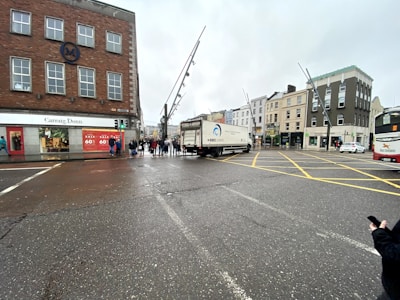 A city street intersection with several buildings, including a store with a large sale sign advertising a 60% discount. A truck is taking a turn in the middle of the intersection, and there are pedestrians on the sidewalk waiting to cross. A bus is visible on the right side of the image.
