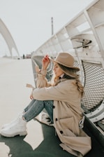 woman in brown coat and blue denim jeans sitting on gray metal bench during daytime
