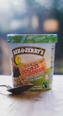 A container of Ben & Jerry's Cookies on Cookie Dough Non-Dairy vegan ice cream placed on a wooden surface. The container features a green lid with the word 'VEGAN' printed repeatedly. The label shows images of cookies and chocolate chunks and includes Fairtrade and non-dairy certifications. A spoon is placed in front of the container.