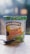A container of Ben & Jerry's Cookies on Cookie Dough Non-Dairy vegan ice cream placed on a wooden surface. The container features a green lid with the word 'VEGAN' printed repeatedly. The label shows images of cookies and chocolate chunks and includes Fairtrade and non-dairy certifications. A spoon is placed in front of the container.