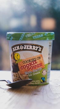 A container of Ben & Jerry's Cookies on Cookie Dough Non-Dairy vegan ice cream placed on a wooden surface. The container features a green lid with the word 'VEGAN' printed repeatedly. The label shows images of cookies and chocolate chunks and includes Fairtrade and non-dairy certifications. A spoon is placed in front of the container.